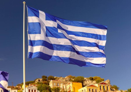 Vibrante bandera griega ondeando sobre coloridas casas de la encantadora ciudad griega, Isla Symi, Grecia