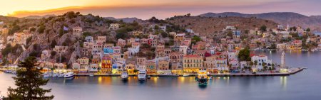 Panoramic view of Symi Harbor at sunset with illuminated buildings and boats, Greece