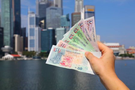 Mano sosteniendo dólares de Singapur papel moneda con horizonte de la ciudad de Singapur en segundo plano.