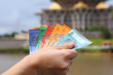 Hand hält malaysisches Ringgit-Papiergeld mit der Skyline von Kuching im Hintergrund.