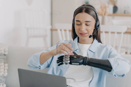 Photo for Female student with disability wearing headset learning at laptop at home. Disabled girl setting, touching her modern bionic prosthetic arm before work or distant education on computer. - Royalty Free Image