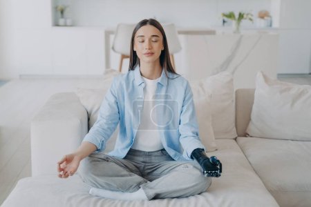 Photo for Calm disabled girl practicing yoga, sitting in lotus pose while meditation. Young female does mudra gesture with bionic prosthesis arm on sofa at home. Healthy lifestyle of people with disabilities. - Royalty Free Image