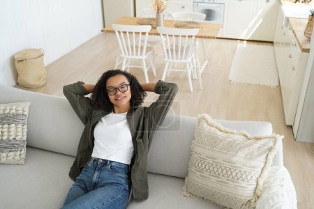 Photo for Relaxed biracial young girl in glasses rest, dreaming, sitting on couch in cozy living room at home. Calm woman relaxing on comfortable sofa with hands behind head, enjoying lazy break. - Royalty Free Image