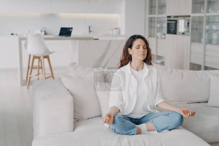 Photo for Hispanic woman makes mudra gesture sitting in lotus posture on comfortable sofa at home. Female practicing yoga, meditates, doing breathing exercises, enjoying fresh air. Wellness, healthy lifestyle. - Royalty Free Image