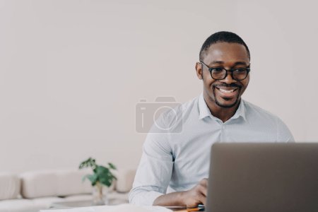 Foto de Sonriente hombre afroamericano desarrollador de software con gafas que trabajan en el proyecto en línea en el ordenador portátil. Programador negro feliz del individuo que crea la aplicación moderna o el email de la lectura con buenas noticias. - Imagen libre de derechos