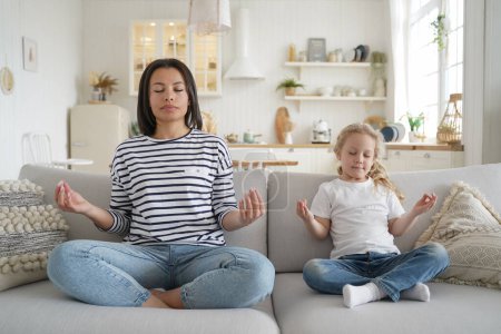Photo for Young mom or babysitter and adopted little daughter practicing yoga together, sitting on sofa at home. Calm mother and kid girl relaxing together in lotus position on couch. Healthy lifestyle concept. - Royalty Free Image