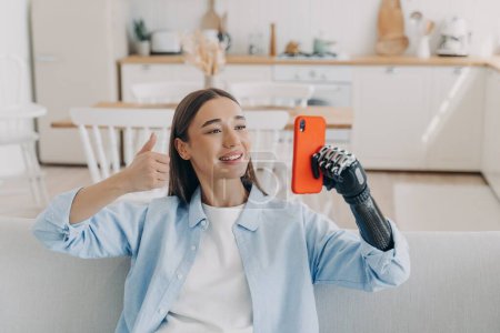 Handicapped confident woman has video call on smartphone. Happy european girl is holding the phone with bionic artificial arm. Attractive handicapped woman at home. Equality and life quality concept.