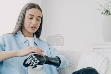 Photo for Handicapped girl is exploring her bionic arm and pressing buttons. Young european woman with cyber hand at home. Modern bionic prosthesis. Futuristic technology of artificial limbs. - Royalty Free Image