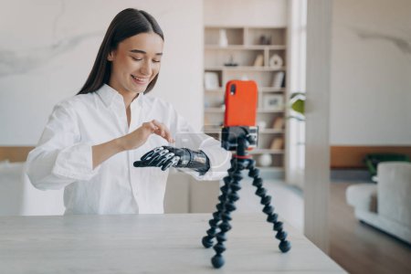 Photo for Happy european handicapped woman is filming on phone camera. Amputee is taking video how to use robotic cyber arm. Pretty disabled girl is blogger. Rehabilitation and wellbeing concept. - Royalty Free Image