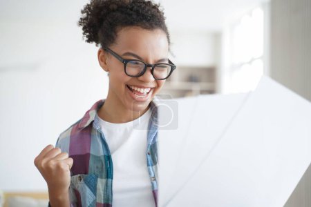 Photo for Excited spanish schoolgirl is winner, holding letter. Pupil is accepted to college. Afro teenage girl is celebrating her victory. Exam is passed successfully. Remote study, distance learning at home. - Royalty Free Image