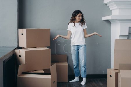 Photo for Puzzled female tenant shrugging looking around, suffers stress with packing things and leaving rented apartment. Tired girl standing with cardboard boxes in vacated house. Hard relocation day. - Royalty Free Image