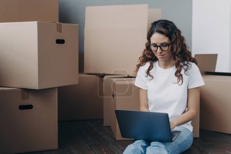 Hispanic woman with laptop searching for mover for relocation online sitting on floor with cardboard boxes. Focused female in glasses reviewing arrangements with moving company on computer.