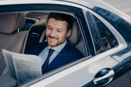 Photo for Handsome young banker in smart formal tuxedo suit reads newspaper with smile, while sitting on passenger seat in expensive car, goes to corporate executive meeting in new office - Royalty Free Image