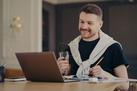 Photo for Happy man freelancer or enterpruer in casual wear sitting at desk in front of opened laptop computer and surfing internet, holding glass of water and working online from home. Freelance concept - Royalty Free Image