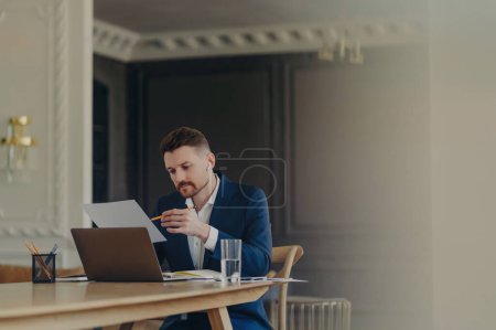 Photo for Handsome business executive in dark blue suit sitting at his work desk in light colored office working on data presented on A4 sheet, holding pencil while being serious and concentrated - Royalty Free Image