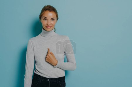 Photo for Satisfied pleased woman points at herself being chosen receives great news dressed in turtleneck and jeans isolated over blue background with copy space for your advertisement. Do you mean me - Royalty Free Image