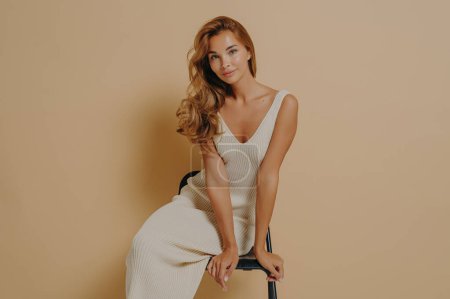 Photo for Women beauty. Adorable skinny young female model wears light knitted dress posing seated on chair leaning forward with lovely smile looking at camera, isolated on beige studio background - Royalty Free Image