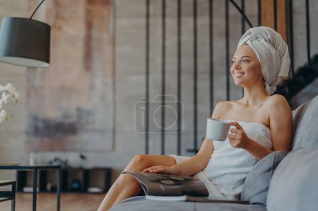 Photo for Photo of relaxed smiling woman enjoys drinking tea, reads magazine and poses on comfortable sofa in living room, enjoys spare time during day off, wears minimal makeup, has well cared healthy skin - Royalty Free Image