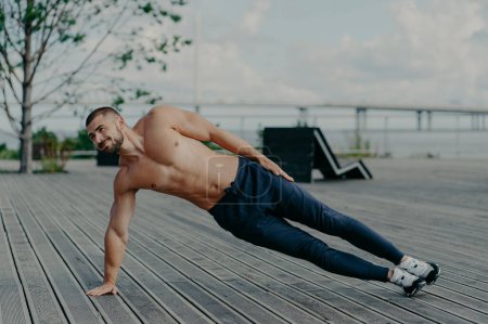 Photo for Sporty bodybuilder stands in side plank, exercises outdoor and wears sport clothes, leads healthy lifestyle, has fit body and muscles. Outdoor shot of determined sportsman does health activities - Royalty Free Image