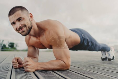 Photo for Workout and healthy lifestyle concept. Pleased muscular bearded young man stands in plank pose, makes sport training outdoors, looks with determined expression, concentrated on getting better - Royalty Free Image