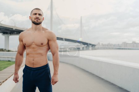 Photo for Handsome muscular man with naked torso has outdoor fitness workout looks into distance, has sexy body, dressed in sports trousers stands outside near river bridge. Athletic guy runs in morning - Royalty Free Image