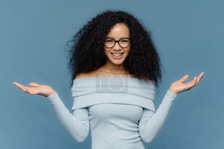 Photo for Waist up shot of happy young crisp Afro American woman raises both hands, pretends holding something, wears blue sweater, has naked shoulders, optical glasses, isolated over blue background. - Royalty Free Image
