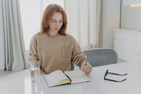 Photo for Indoor shot of serious redhead young woman makes notes in notepad makes list to do plans workday manager time sits at white desktop with glass of water spectacles against cozy domestic interior - Royalty Free Image