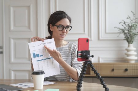 Photo for Young hispanic woman real estate advisor talking with client online about monthly mortgage payment, sitting in front of mobile phone on tripod, female loan officer working remotely from home - Royalty Free Image
