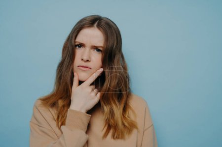 Photo for Puzzled young girl with thick wavy ombre hair demonstrating concern and interest while holding hand at chin wearing casual light brown sweater and standing against blue studio background - Royalty Free Image