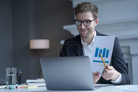 Photo for Confident young male company CEO in formal wear holds online shareholder remote meeting tells and shows charts of income pointing with pencil on graphs in his hand positively smiling on laptop camera - Royalty Free Image