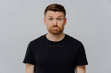Foto de Young redhead man looking displeased, seeing something disgusting, dissatisfied guy in black tshirt looking at camera with pouted lips while standing over gray background. Negative feelings concept - Imagen libre de derechos