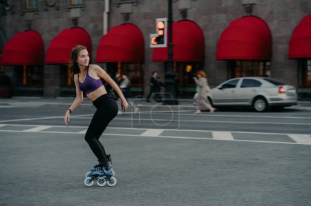 Foto de Full length shot of active slim young woman dressed in sportsclothes rides on rollers to strengthen arms and legs muscles improves balance agility and coordination has good mood burns calories - Imagen libre de derechos
