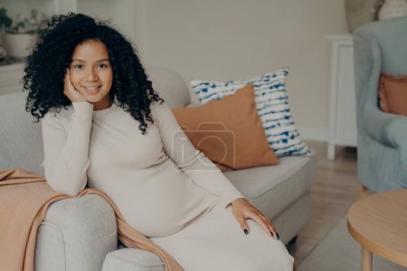 Téléchargez les photos : Belle femme africaine enceinte heureuse assise dans une pose détendue tout en étant enceinte, la tête reposée sur le côté du canapé confortable dans le salon, souriant et profitant de son temps libre à la maison - en image libre de droit