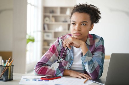 Photo for Pensive young student girl sitting at table with laptop looks into distance, studying at home. Thoughtful puzzled mixed race schoolgirl ponders solution, preparing for exam, makes homework task. - Royalty Free Image