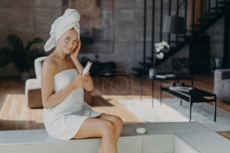 Photo for Pleased healthy woman applies body lotion and face cream, nourishes skin, wrapped in bath towel, smiles gently, poses against domestic interior, undergoes beauty treatments. Dermatology concept - Royalty Free Image