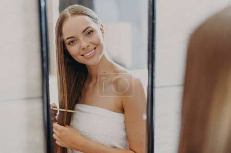 Photo for Happy beautiful young woman looks at her reflection in mirror, has well cared long hair, uses hair brush, wrapped in bath towel, makes hairstyle. Women, beauty, hairstyling and self care concept - Royalty Free Image
