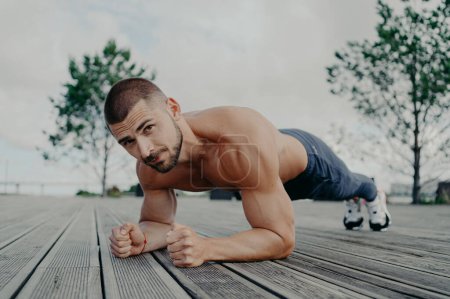 Photo for Motivational bearded man stands in plank pose, trains muscles and has strong body. Sporty adult guy performs push up exercise outdoor. Bodybuilder has abdominal workout. Active lifestyle concept - Royalty Free Image