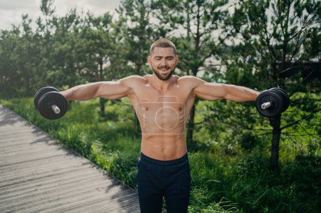 Photo for Fit adult man lifts heavy weight during workout session outdoor, poses with naked torso, has well shaped muscular body, demonstrates his power, lifts barbells. Motivated bodybuilder at street - Royalty Free Image