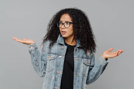 Confused unaware Afro woman with crisp hair, raises hands in bewilderment, looks aside, cannot make decision, wears spectacles and jean jacket, isolated over grey background. What should I do now?