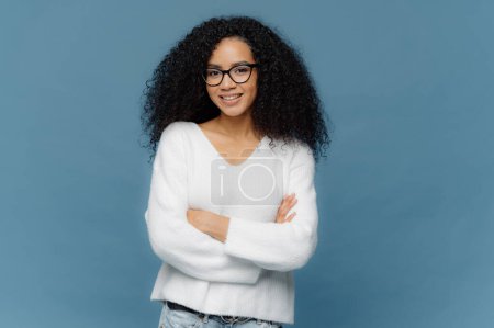 Photo for Portrait of delighted pleasant looking curly female keeps arms folded on chest, has confident facial expression, charmin smile on face, wears white sweater and jeans, stands over blue background. - Royalty Free Image