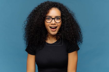 Photo for Pleasantly surprised dark skinned woman keeps jaw dropped, gazes with interest, has curly hair, wears black t shirt, models against blue wall. Afro American lady gazes with shock, has bated breath - Royalty Free Image