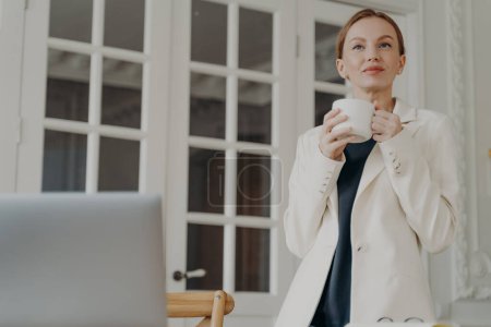 Photo for Elegant business lady in formal wear is working at home or office in evening. Glamorous mid adult businesswoman is dreaming with cup of tea. Attractive european woman is a boss or entrepreneur. - Royalty Free Image