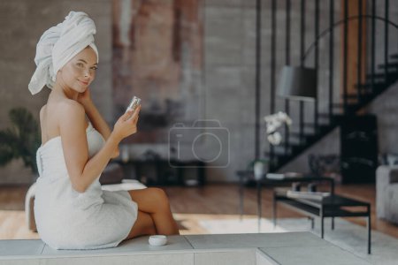 Photo for Indoor shot of good looking woman holds bottle of lotion has well cared smooth skin wears towel on head and around naked body after taking shower, poses against home interior. Beauty concept. - Royalty Free Image