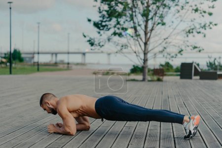 Photo for Horizontal shot of sporty bearded man stands in plank pose, practices yoga outdoor and dressed in active wear, breathes fresh air. Sport, fitness and healthy lifestyle concept. Training workout - Royalty Free Image