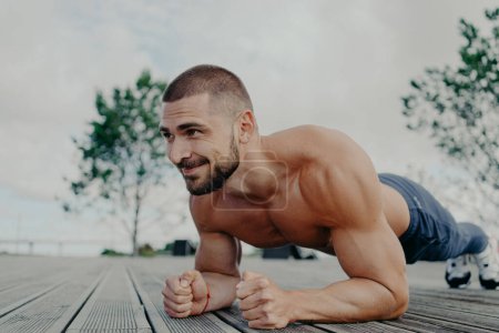 Photo for Athletic guy stands in plank pose to keep fit, leads healthy lifestyle and poses outdoor. Young bearded man in good physical shape does abdominal excersice, has muscular torso. Bodybuilding concept - Royalty Free Image