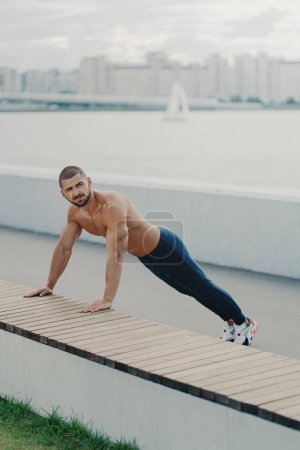 Photo for Outdoor shot of determined muscular man warms up before training stands in plank pose has naked torso muscular body enjoys exercising in open air wears active wear has workout to maintain vitality - Royalty Free Image