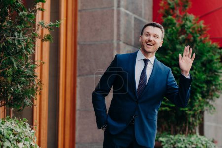 Téléchargez les photos : Greeting concept. Cheerful successful young businessman in formal clothes waves with hands as sees colleague into distance, has happy expression, stands outdoor near restaurant. Hey, I am here! - en image libre de droit