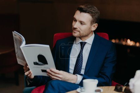 Photo for Horizontal shot of attractive male manager dressed in black suit, white shirt and tie, reads magazine in coffee shop, drinks hot beverage, has serious concetrated look. Business and leisure concept - Royalty Free Image