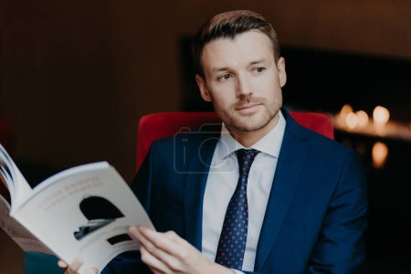 Téléchargez les photos : Pensive successful male employee focused into distnace, holds magazine, dressed in formal clothes, waits for business partner in cafe, thinks about signing contract and collaboration. People and work - en image libre de droit