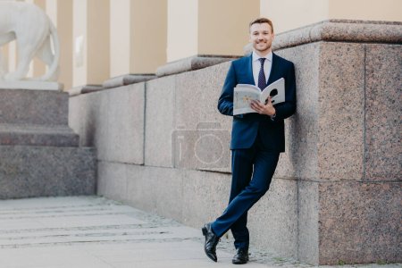Foto de Photo of successful male boss stands crossed legs, holds business magazine, reads necessary information, prepares for presentation in front of colleagues, stands near office building, dressed formally - Imagen libre de derechos